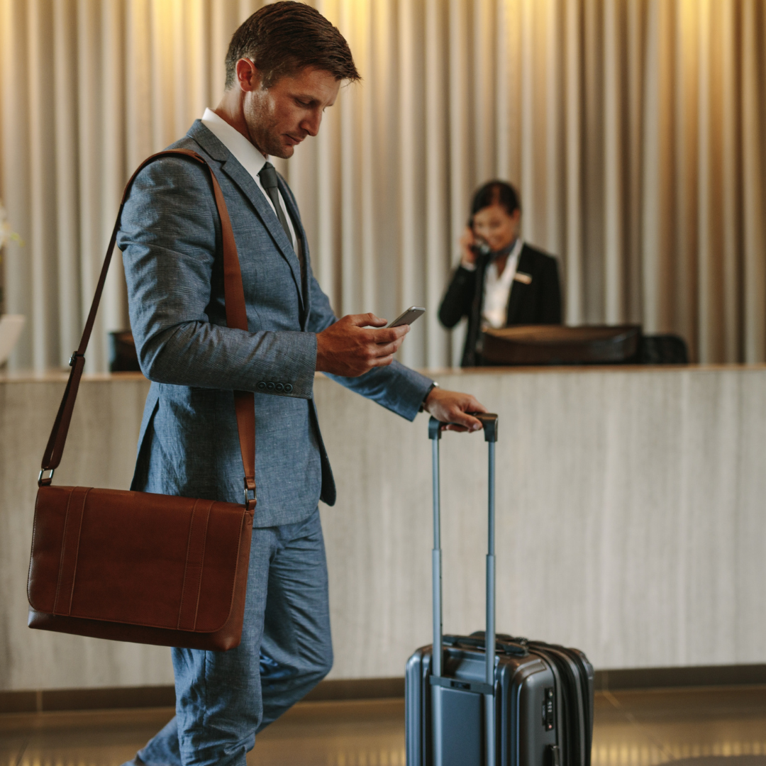 Business traveller holding a work bag and a small suitcase looks at his phone while the hotel receptionist conducts a call in the background